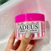 Adeus Cream cellulite and stretch mark cream in a pink and white striped jar held by a hand