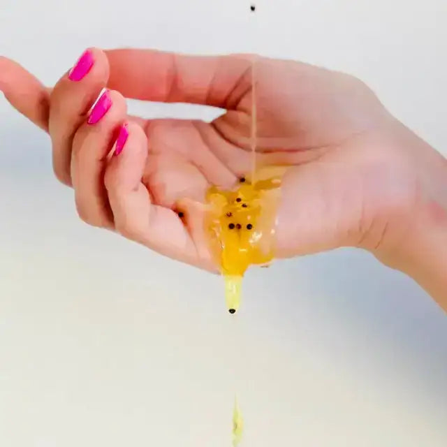 Adeus Banana Skin Brightening Exfoliator – Gentle Body & Face Renewal Scrub, 5.82 oz (165g) - Close-up showing rich yellow-orange texture and exfoliating particles dripping from a hand with pink nails – Buy now at MKPBR.COM – Worldwide Shipping