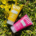 The Adeus Duo Exfoliator Kit, featuring the yellow banana and pink pitaya skin renewal and brightening scrubs, lying on lush green grass with delicate yellow flowers in a natural outdoor setting.– Buy now at MKPBR.COM – Worldwide Shipping
