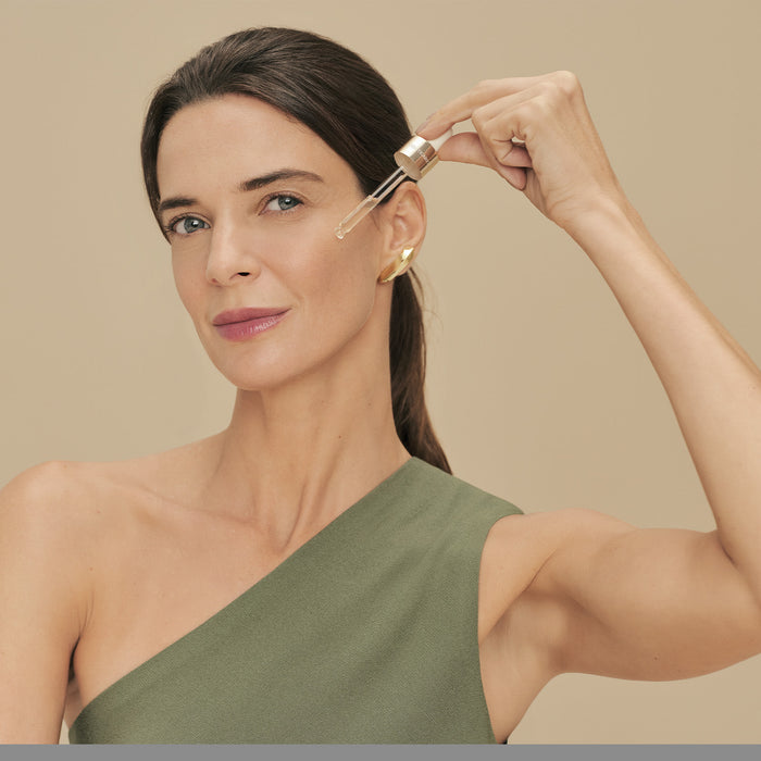 Model applying a drop of Boticário Botik Peptídeos Pró-Elastina Facial Serum to her cheek with the glass dropper, Brazilian skincare routine – Buy now at MKPBR.COM – Worldwide Shipping