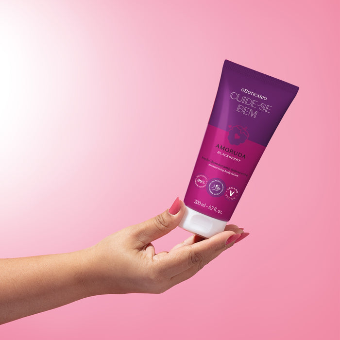 Hand holding the O Boticário Cuide-se Bem Amoruda moisturizing body lotion tube against a soft pink backdrop – Buy now at MKPBR.COM – Worldwide Shipping