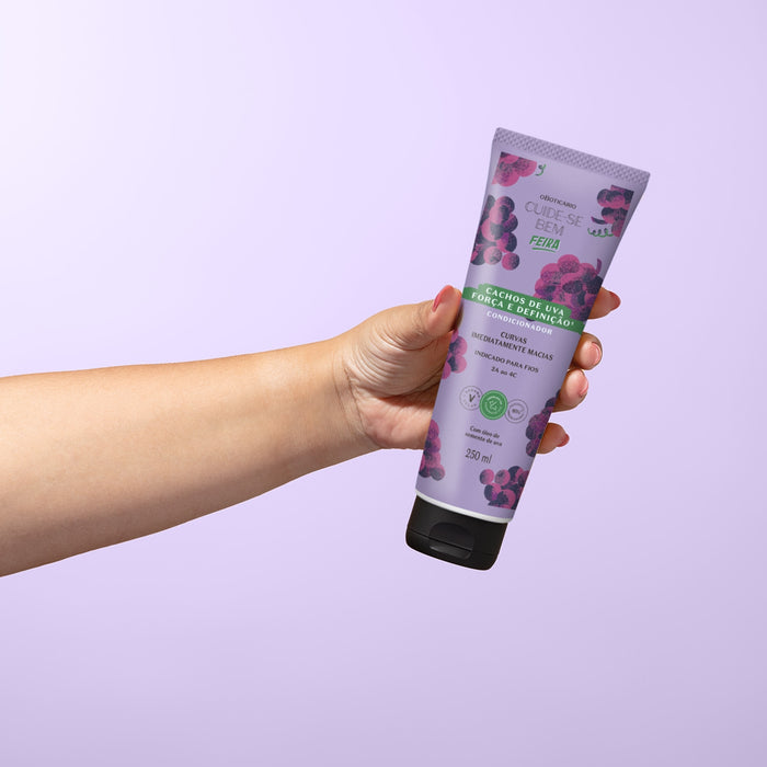 Hand holding the O Boticário Cachos de Uva grape curl conditioner tube against a lilac background showing the 250 ml size for curls 2A to 4C – Buy now at MKPBR.COM – Worldwide Shipping