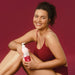 Model seated on a burgundy background holding the 400 ml Boticário Cuide-se Bem Cereja de Fases body lotion bottle against her leg – Buy now at MKPBR.COM – Worldwide Shipping