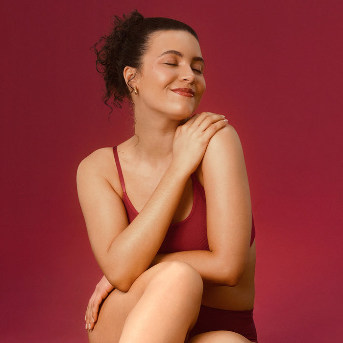 Smiling model with soft glowing skin resting her hand on her shoulder against a burgundy backdrop, styled for Boticário Cuide-se Bem Cereja de Fases body lotion – Buy now at MKPBR.COM – Worldwide Shipping