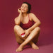 Smiling model in red seated on a red background applying Boticário Cuide-se Bem Cereja de Fases relaxing body cream to her leg – Buy now at MKPBR.COM – Worldwide Shipping