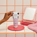 Hand reaching for the O Boticário Cuide-se Bem Deleite body lotion on a pink tiled bathroom counter beside a mirror and towel, everyday Brazilian skincare ritual – Buy now at MKPBR.COM – Worldwide Shipping
