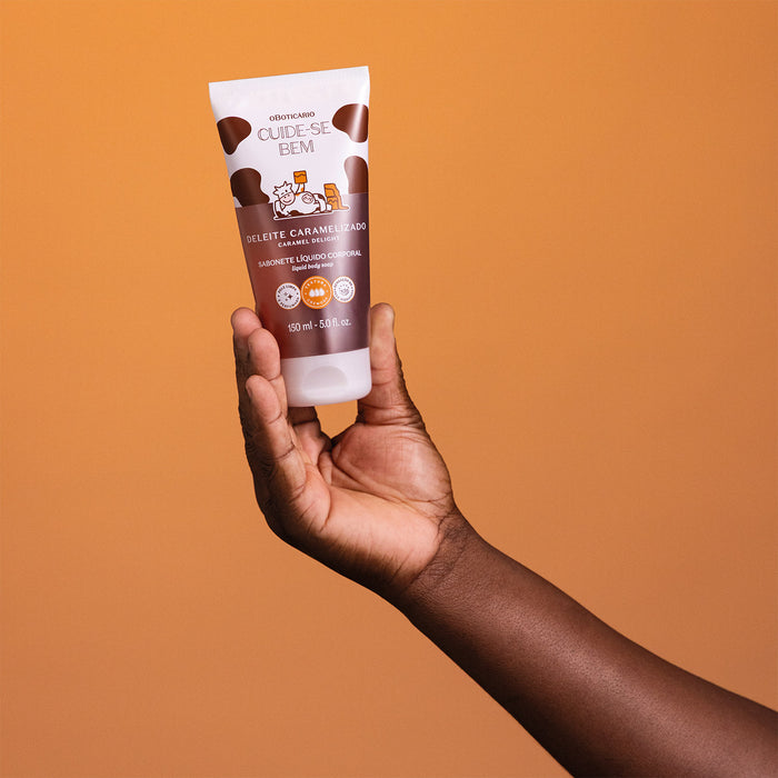 Hand holding the Boticário Cuide-se Bem Deleite Caramelizado Body Wash 150 ml tube against an amber background to show its travel friendly size – Buy now at MKPBR.COM – Worldwide Shipping