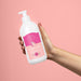 Hand holding the Boticário Cuide-se Bem Doçura na Pessegura 400 ml deodorant body lotion bottle against a pink background for scale – Buy now at MKPBR.COM – Worldwide Shipping