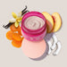 Open jar of Boticário Doçura na Pessegura exfoliating body cream surrounded by peach slices, coconut pieces, vanilla and dried apricots – Buy now at MKPBR.COM – Worldwide Shipping