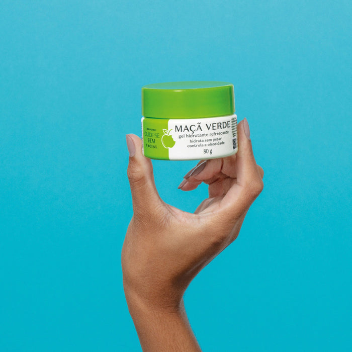 Hand holding the closed 80 g jar of Boticário Cuide-se Bem Feira Maçã Verde facial gel moisturizer against a blue background – Buy now at MKPBR.COM – Worldwide Shipping