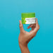 Hand holding the closed 80 g jar of Boticário Cuide-se Bem Feira Maçã Verde facial gel moisturizer against a blue background – Buy now at MKPBR.COM – Worldwide Shipping