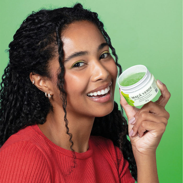 Smiling model holding the open jar of Boticário Cuide-se Bem Feira green apple facial gel moisturizer against a green background – Buy now at MKPBR.COM – Worldwide Shipping