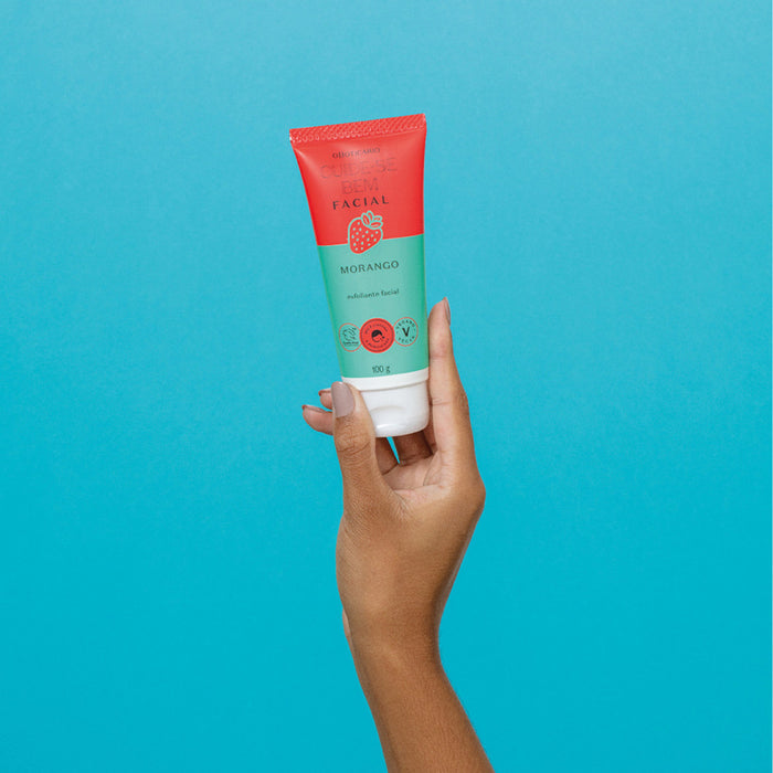 Hand holding the 100 g Boticário Cuide-se Bem Feira Morango strawberry facial scrub tube against a turquoise background – Buy now at MKPBR.COM – Worldwide Shipping