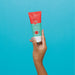 Hand holding the 100 g Boticário Cuide-se Bem Feira Morango strawberry facial scrub tube against a turquoise background – Buy now at MKPBR.COM – Worldwide Shipping