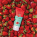 Boticário Cuide-se Bem Feira Morango facial scrub tube lying on a bed of fresh red strawberries – Buy now at MKPBR.COM – Worldwide Shipping