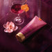 Boticário Glamour Intense Body Lotion tube styled on purple marble with orange liqueur cocktail and magnolia flower showing the sweet floral fragrance notes – Buy now at MKPBR.COM – Worldwide Shipping