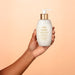 Hand holding the Boticário Lily Acetinado Satin Conditioner 250 ml bottle against a peach backdrop, showing its compact pump size – Buy now at MKPBR.COM – Worldwide Shipping
