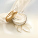 Open jar of Boticário Lily Cashmere Satin Body Cream showing the swirled velvety cream beside a cream silk scarf and golden lid – Buy now at MKPBR.COM – Worldwide Shipping