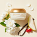 Boticário Lily Cashmere body cream jar surrounded by white floral petals, vanilla pods, pear and red berries showing the Floral Woody fragrance notes – Buy now at MKPBR.COM – Worldwide Shipping