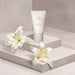 O Boticário Lily Satin Hand Cream 50 g tube on a stone pedestal beside fresh white lily blooms, Brazilian floral hand care – Buy now at MKPBR.COM – Worldwide Shipping
