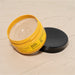 Open jar of Boticário Nativa SPA Ameixa Dourada exfoliating body cream lying beside its black lid on a beige textured backdrop, scrub cream visible inside – Buy now at MKPBR.COM – Worldwide Shipping