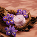 Open jar of Boticário Liz D'Essence body cream showing pink cream, styled with purple iris flowers and driftwood – Buy now at MKPBR.COM – Worldwide Shipping