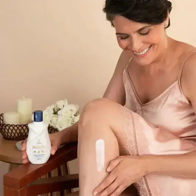 Smiling mature woman seated while applying white Paixão Inspiradora lotion to her leg, bottle and white roses visible in background. Buy now at MKPBR.COM – Worldwide Shipping