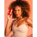 Smiling woman with curly brown hair holding a red bottle of Muriel Rosehip & Collagen Body Oil, promoting glowing skin. Buy now at MKPBR.COM – Worldwide Shipping