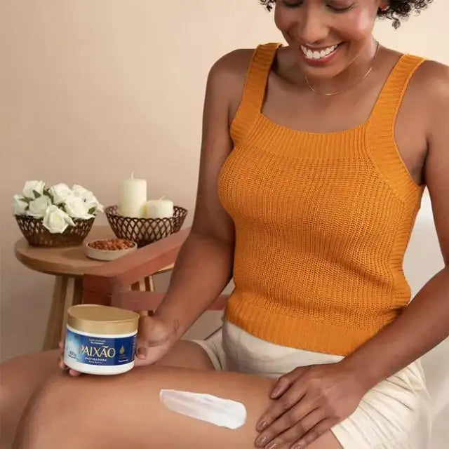 Smiling woman with dark skin applying Paixão Inspiradora Rosas Brancas body cream to her leg — Buy now at MKPBR.COM – Worldwide Shipping