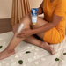 Woman applying Paixão Inspiradora Rosas Brancas ultra-creamy deo hydrating body lotion to her leg; almond oil for hydrated, velvety skin with white rose petals around. Buy now at MKPBR.COM – Worldwide Shipping