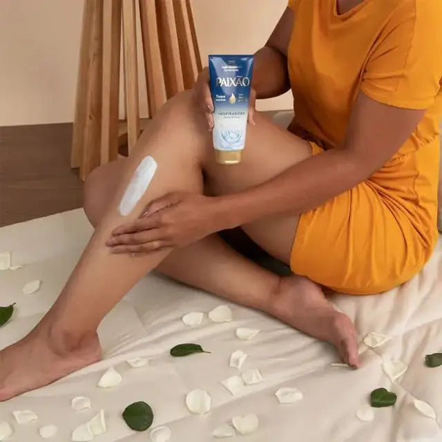 Woman applying Paixão Inspiradora Rosas Brancas ultra-creamy deo hydrating body lotion to her leg; almond oil for hydrated, velvety skin with white rose petals around. Buy now at MKPBR.COM – Worldwide Shipping