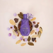 Paixão Irresistível Flor de Lis – purple lotion bottle centered in a ceramic bowl filled with almonds, surrounded by dried petals on a light beige surface – Buy now at MKPBR.COM – Worldwide Shipping