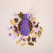 Paixão Irresistível Flor de Lis – purple lotion bottle centered in a ceramic bowl filled with almonds, surrounded by dried petals on a light beige surface – Buy now at MKPBR.COM – Worldwide Shipping