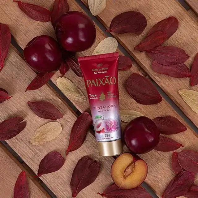 A tube of Paixão Tentadora Ruby Plum hand cream lies on a wooden surface, surrounded by several whole fresh plums, one cut plum revealing its yellow interior, and scattered dark red and light-colored autumn leaves – Buy now at MKPBR.COM – Worldwide Shipping