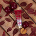 A tube of Paixão Tentadora Ruby Plum hand cream lies on a wooden surface, surrounded by several whole fresh plums, one cut plum revealing its yellow interior, and scattered dark red and light-colored autumn leaves – Buy now at MKPBR.COM – Worldwide Shipping