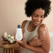 Smiling young Black woman holding Paixão Inspiradora Rosas Brancas lotion and applying white lotion to her arm, with candles and flowers behind. Buy now at MKPBR.COM – Worldwide Shipping